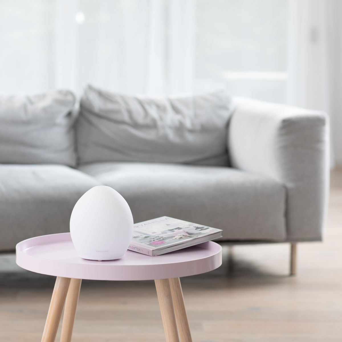 A modern aroma diffuser sits on a small table in front of a sofa in a bright living room.